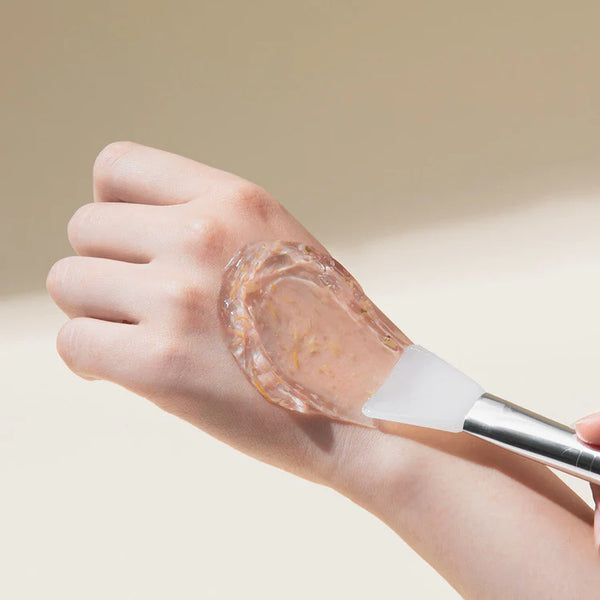 Application of MARY & MAY Calendula Peptide Ageless Sleeping Mask on the back of a hand with a spatula, demonstrating the product's texture and consistency.