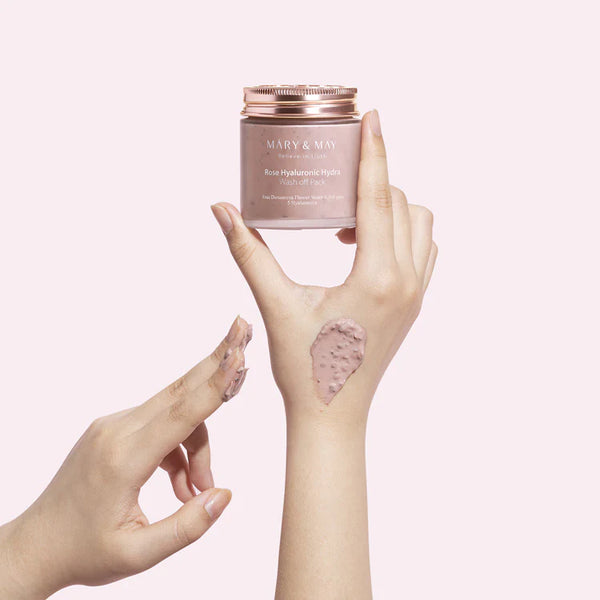 Hand delicately holding the MARY & MAY Rose Hyaluronic Hydra Wash Off Pack jar with rose-infused clay product applied to the wrist.